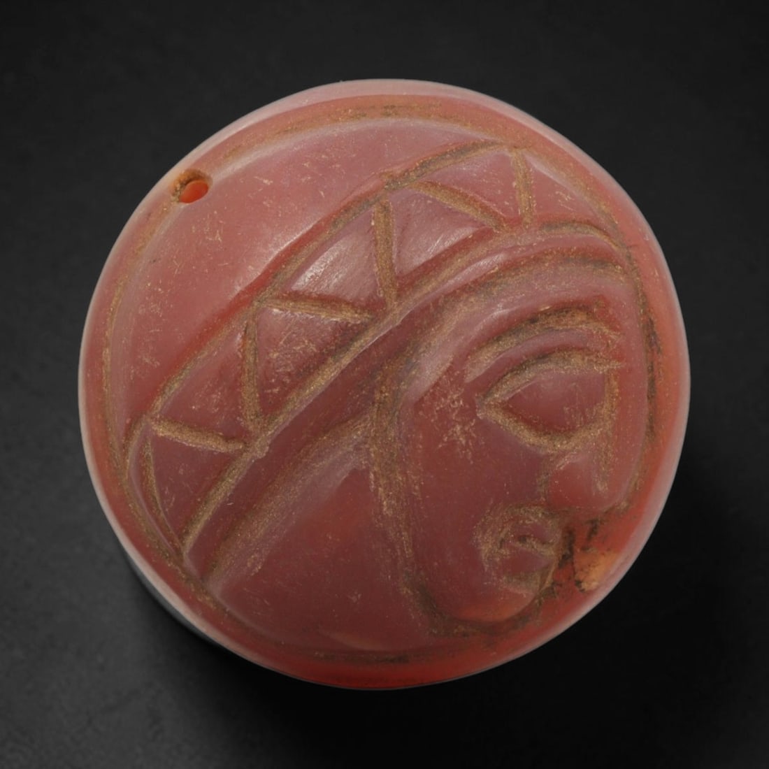 Ancient Carnelian Amulet - Near Eastern Bust Pendant, 44ZX: Carved from carnelian, the amulet is a domed circular disc with a drilled suspension hole at the edge, crafted in the ancient Near Eastern style. The front surface bears a raised relief portrait of a