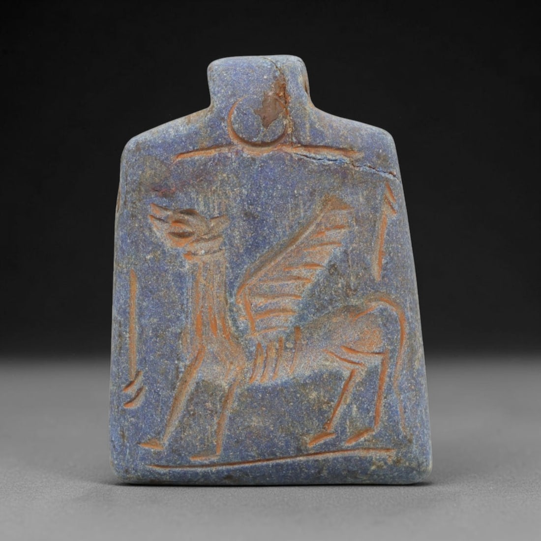 Ancient Lapis Lazuli Amulet - Near Eastern Winged Camel Figure, 49ZX: Carved from lapis lazuli, this amulet features a relief depiction of a winged camel, a mythical hybrid creature, rendered in the stylized glyptic tradition of the ancient Near East. Incised details de