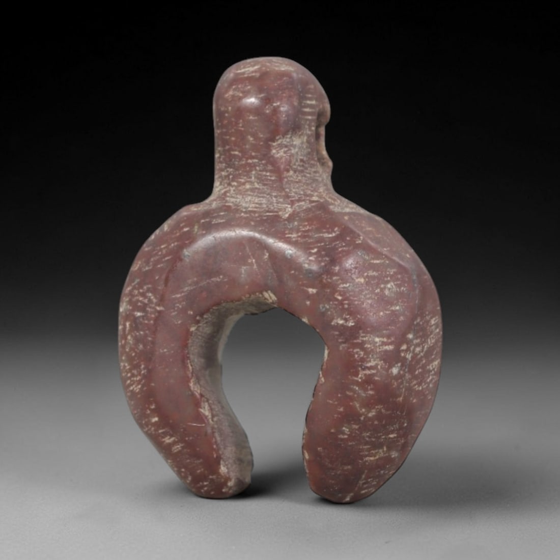 Ancient Stone Amulet - Near Eastern Ritual Figurine, 38AP: This artifact is carved from deep red stone with a weathered, earthy patina and subtle mineral inclusions, exhibiting a stylized, symmetrical U-shaped form consistent with ancient Near Eastern stonewo