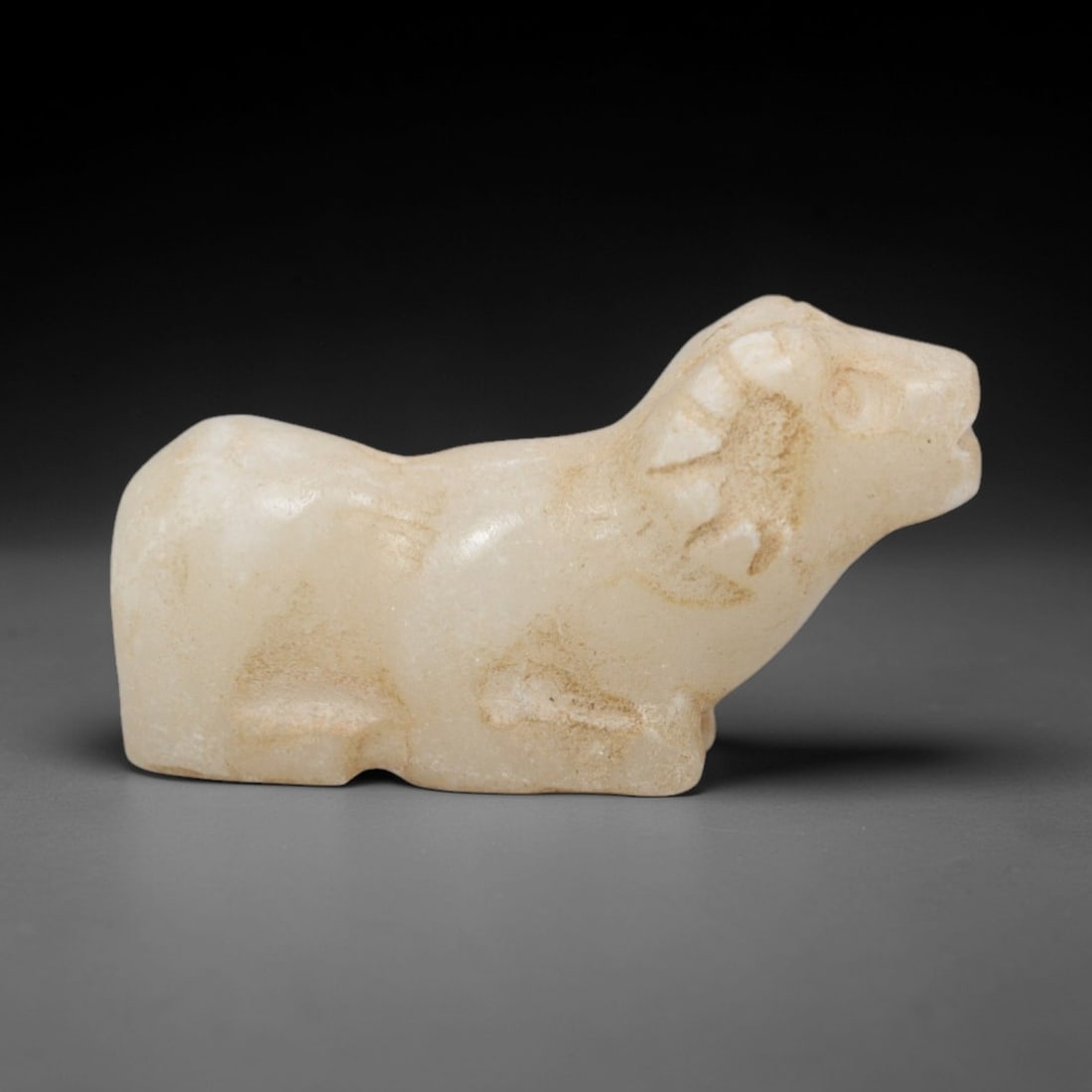 Ancient Marble Ram Amulet - Near Eastern Figurine, 93XR: Crafted from pale marble, this small amulet depicts a recumbent ram in a stylized form. The compact body is rounded, with incised details defining the curved horns, facial contours, and folded limbs,
