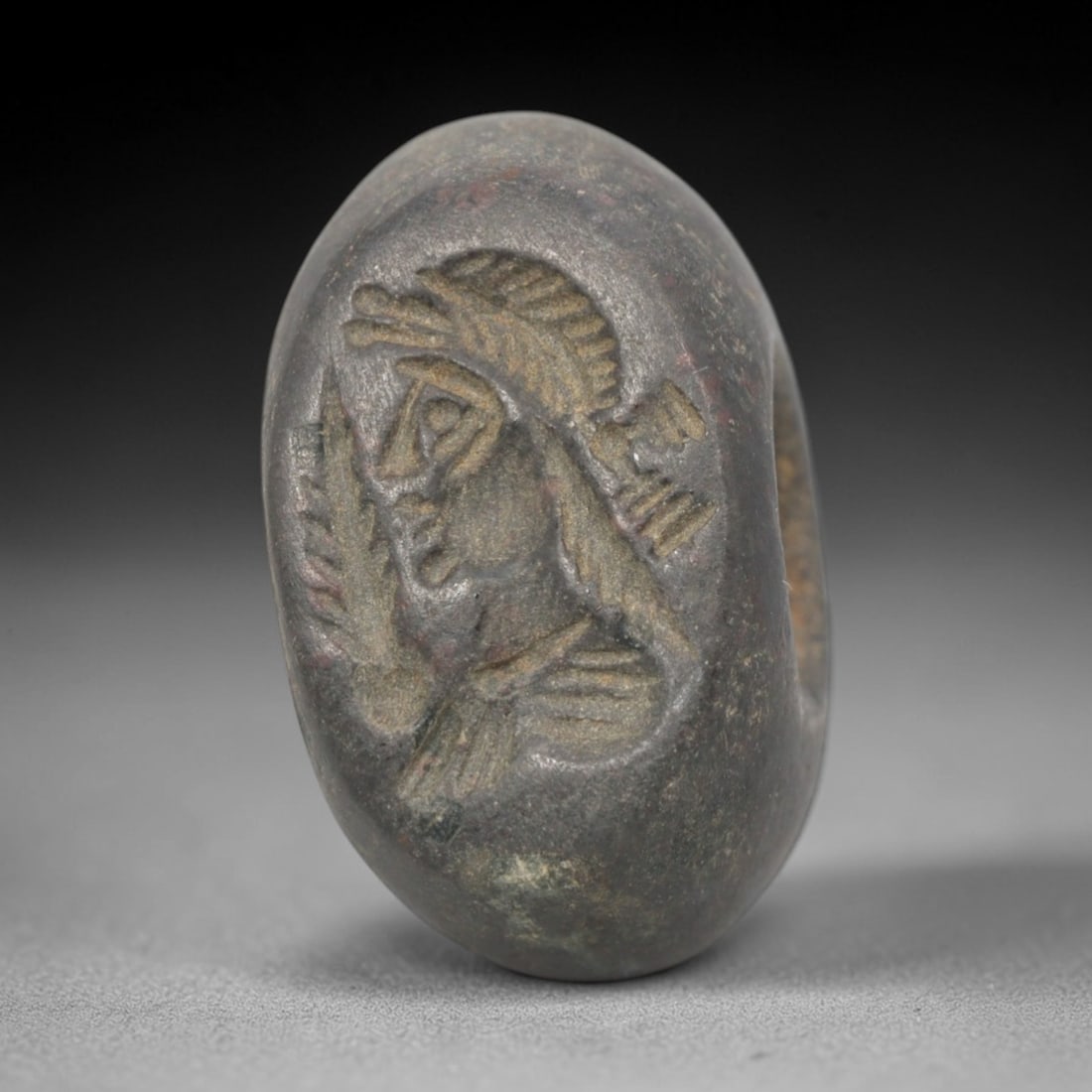 Ancient Stone Hemispherical Stamp Seal - Near Eastern Winged Anthropomorphic Figure, 82ZS: Crafted from dense, dark gray stone, this hemispherical stamp seal bears incised carving of a winged anthropomorphic figure, depicted with a stylized headdress, defined facial features, and outstretch