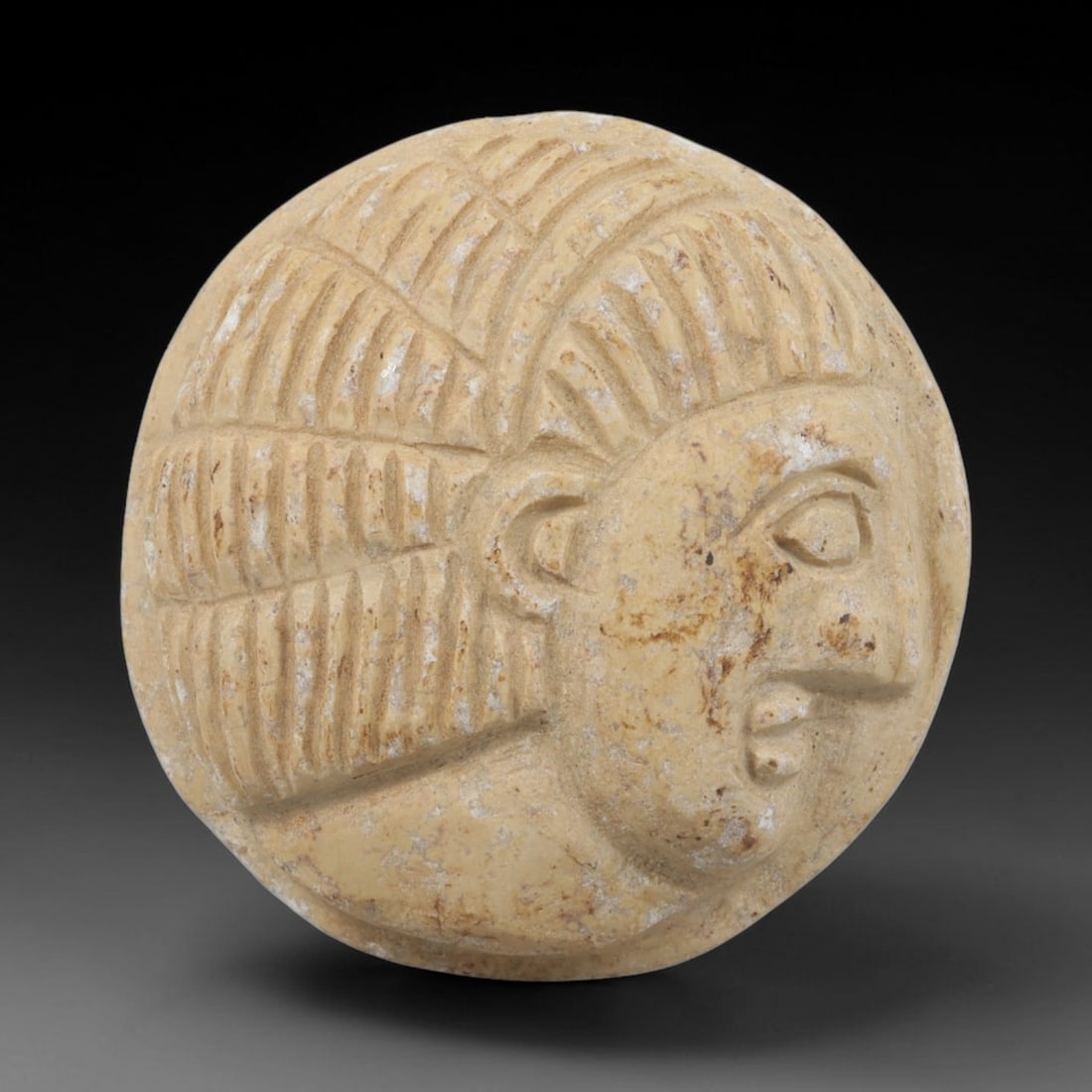 Ancient Near Eastern Stone Amulet - Anthropomorphic Bust Plaque, 80FY: Carved from a fine-grained, creamy-beige stone, this amulet is a circular plaque crafted in the ancient Levantine style, featuring a raised relief portrait of a human bust in profile. The surface bear