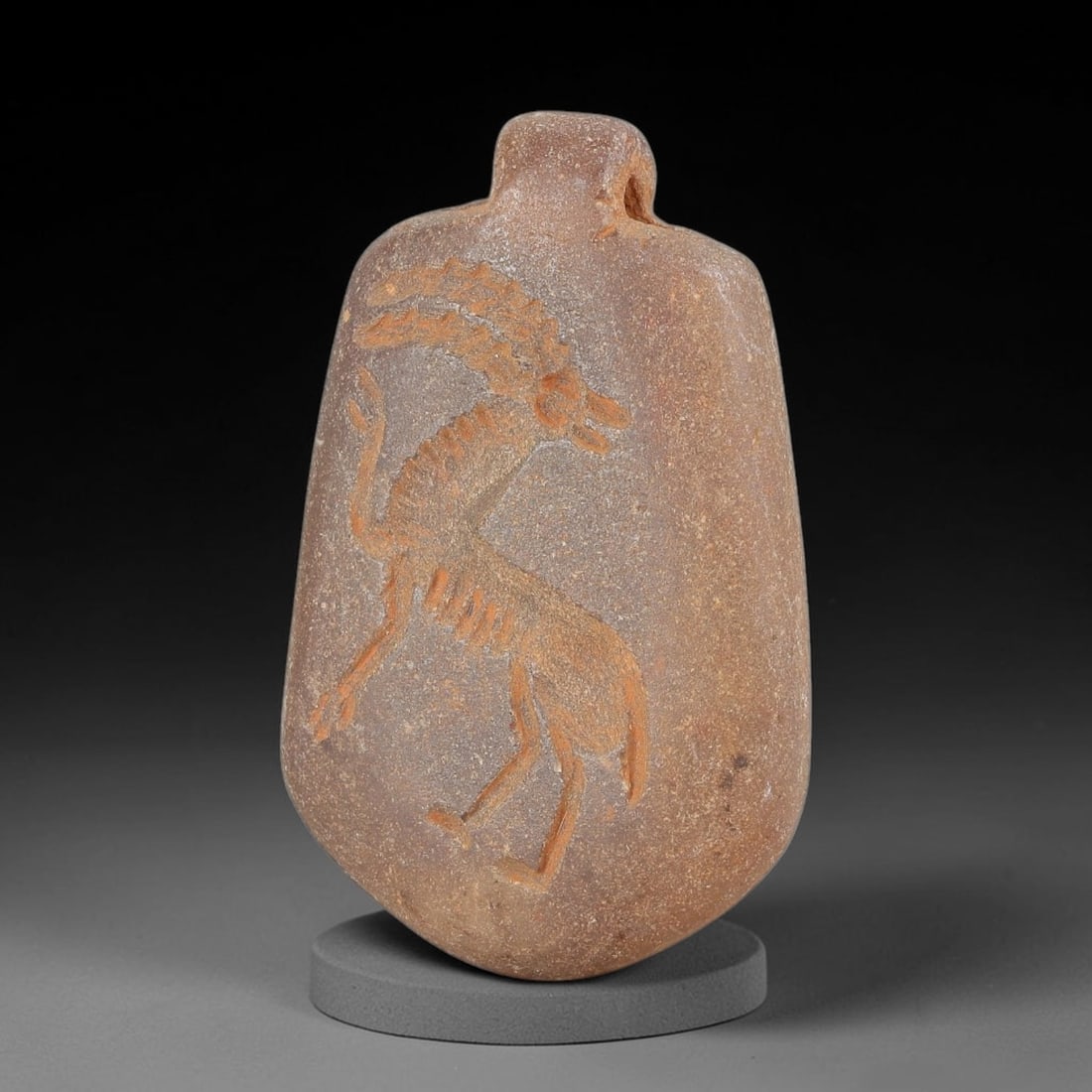 Ancient Stone Ibex Amulet - Near Eastern Carved Pendant, 29BA: Carved from hardstone, the amulet takes the form of a flattened oval pendant with a drilled suspension lug at the top for wear. The surface features a relief-carved stylized ibex (wild goat) in a dyna
