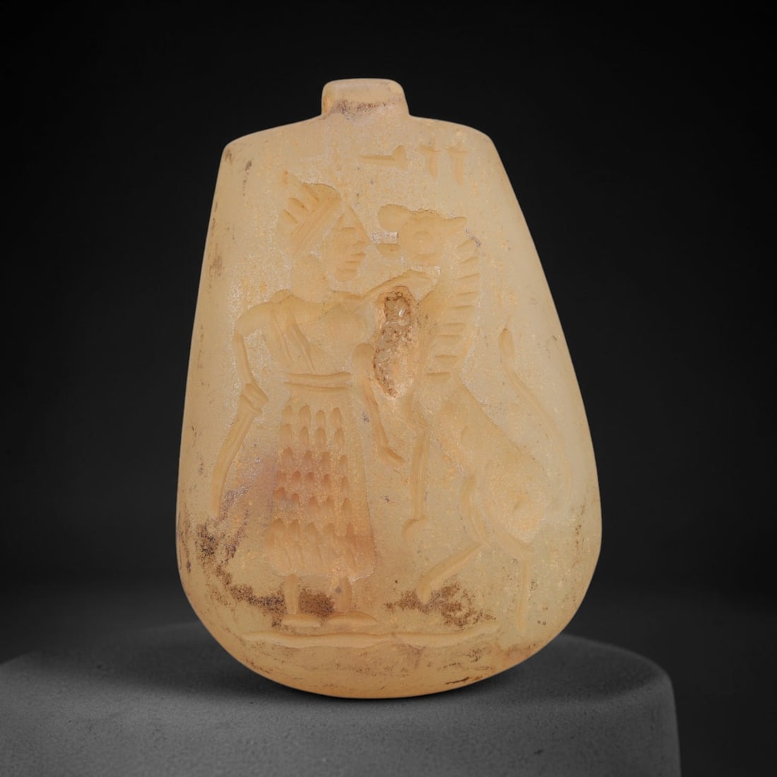 Ancient Chalcedony Amulet - Near Eastern Human & Griffin Figure, 75ZT: Carved from chalcedony, this amulet features a relief scene depicting a standing human figure confronting a rampant griffin, rendered in the stylized glyptic tradition of the ancient Near East. The hu