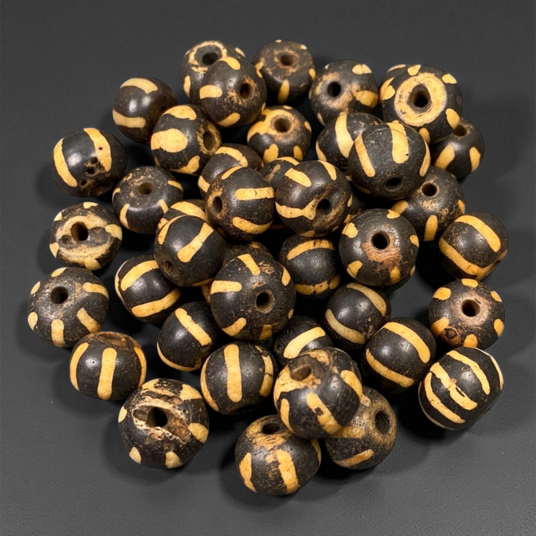 A Group of Burmese Pumtek Wood Beads, 19th-20th Century, 92FS: The group comprises forty-three spherical beads, crafted from wood with a dark treated surface and inlaid light tan geometric motifs. The decoration consists primarily of linear bands, stripes, and si