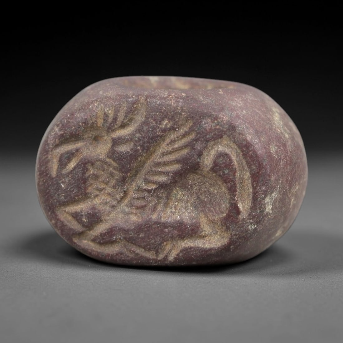 Ancient Stone Hemispherical Stamp Seal - Near Eastern Winged Stag Figure, 14AN: Carved from a dense, dark reddish-brown stone, this hemispherical stamp seal features a flat, oval face incised with a stylized winged stag in profile. The stag is rendered with prominent antlers, fea