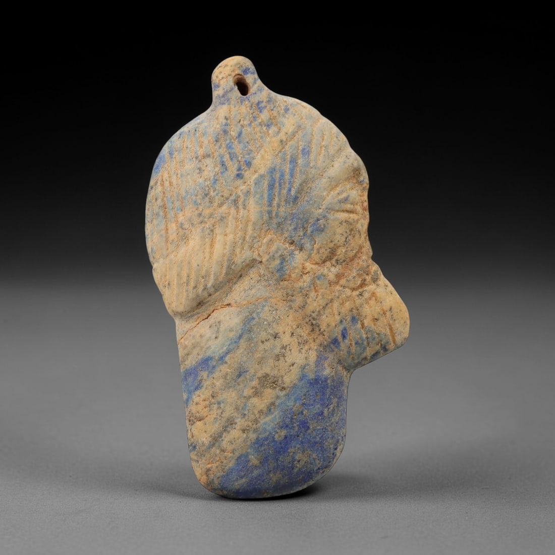 Ancient Lapis Lazuli Noble Head Amulet - Egyptian Pendant, 7AJ: Carved from lapis lazuli stone, this ancient Egyptian noble head amulet pendant features a stylized human head with detailed facial features and a crown-like headdress. The carving exhibits the refine