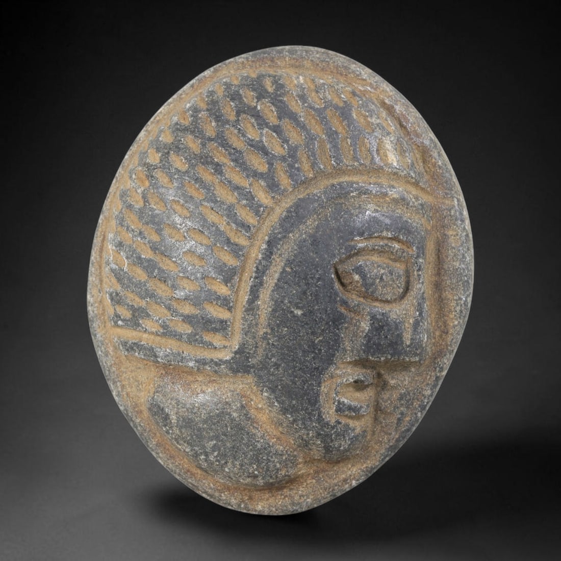 Ancient Stone Head Plaque - Near Eastern Amulet, 41FY: Carved from a dense, dark grey stone, this amulet takes the form of a stylized human head in relief, set within an oval plaque, characteristic of ancient Near Eastern glyptic and amuletic traditions.
