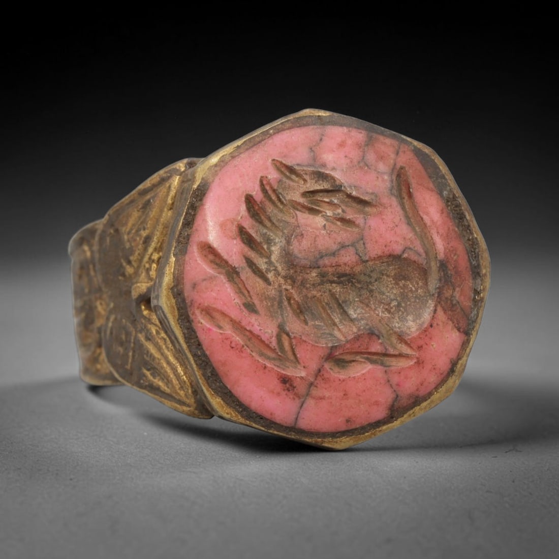 Antique Qajar Dynasty Pink Stone Intaglio Ring - Size 9 US, 2WJ: Crafted from gilt brass with a faceted bezel, this ring features a carved pink stone intaglio at its center, incised with a stylized quadruped motif characteristic of Qajar-era Persian symbolic carvin