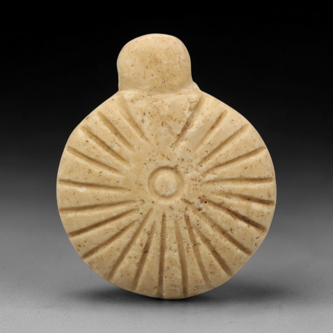 Ancient Limestone Sun Amulet - Near Eastern Pendant, 46ZW: This amulet is carved from pale beige limestone, featuring a circular disc with a raised central boss and radiating incised lines that form a sunburst pattern. A small rounded suspension knob is attac