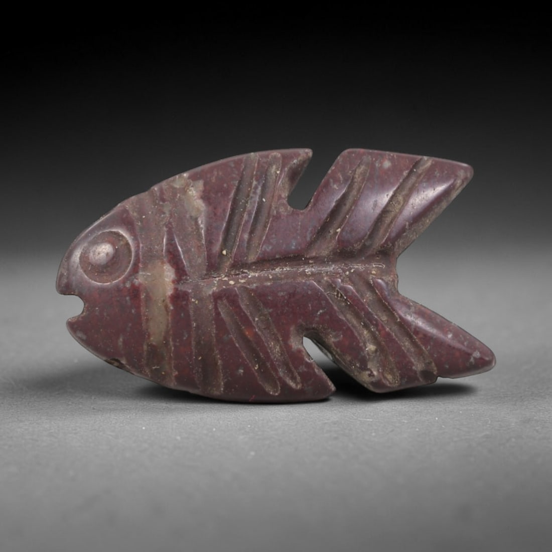 Ancient Stone Fish Amulet - Egyptian Animal Figurine, 86AP: Carved from a dense, reddish stone with natural mineral inclusions and a smooth, weathered patina, this amulet depicts a stylized fish form with a rounded head, a clearly defined eye, and a body taper