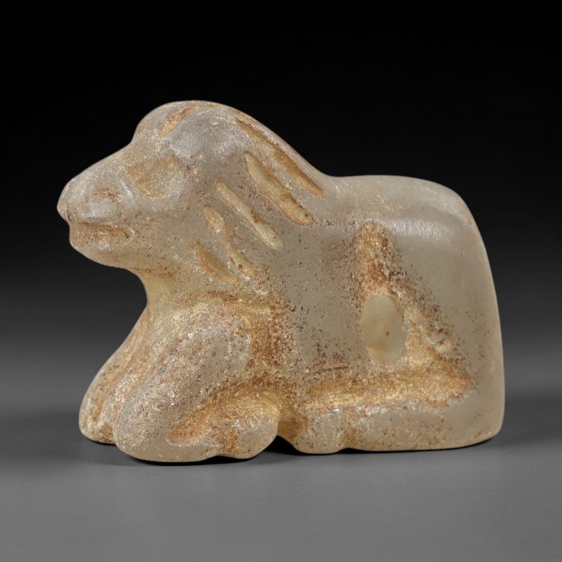 Ancient Glass Feline Amulet - Near Eastern Animal Pendant, 74AK: Carved from translucent pale gray glass, this reclining feline amulet features a stylized head with simplified facial features and linear incised patterns defining its mane. The compact, rounded body