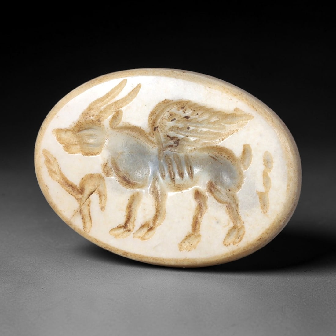 Ancient Stone Winged Bull Intaglio - Near Eastern Amulet, 62AR: This oval intaglio is carved from a pale, layered stone matrix, featuring a high-relief depiction of a winged bull-a hybrid creature with bovine body, curved horns, and feathered avian wings. The crea