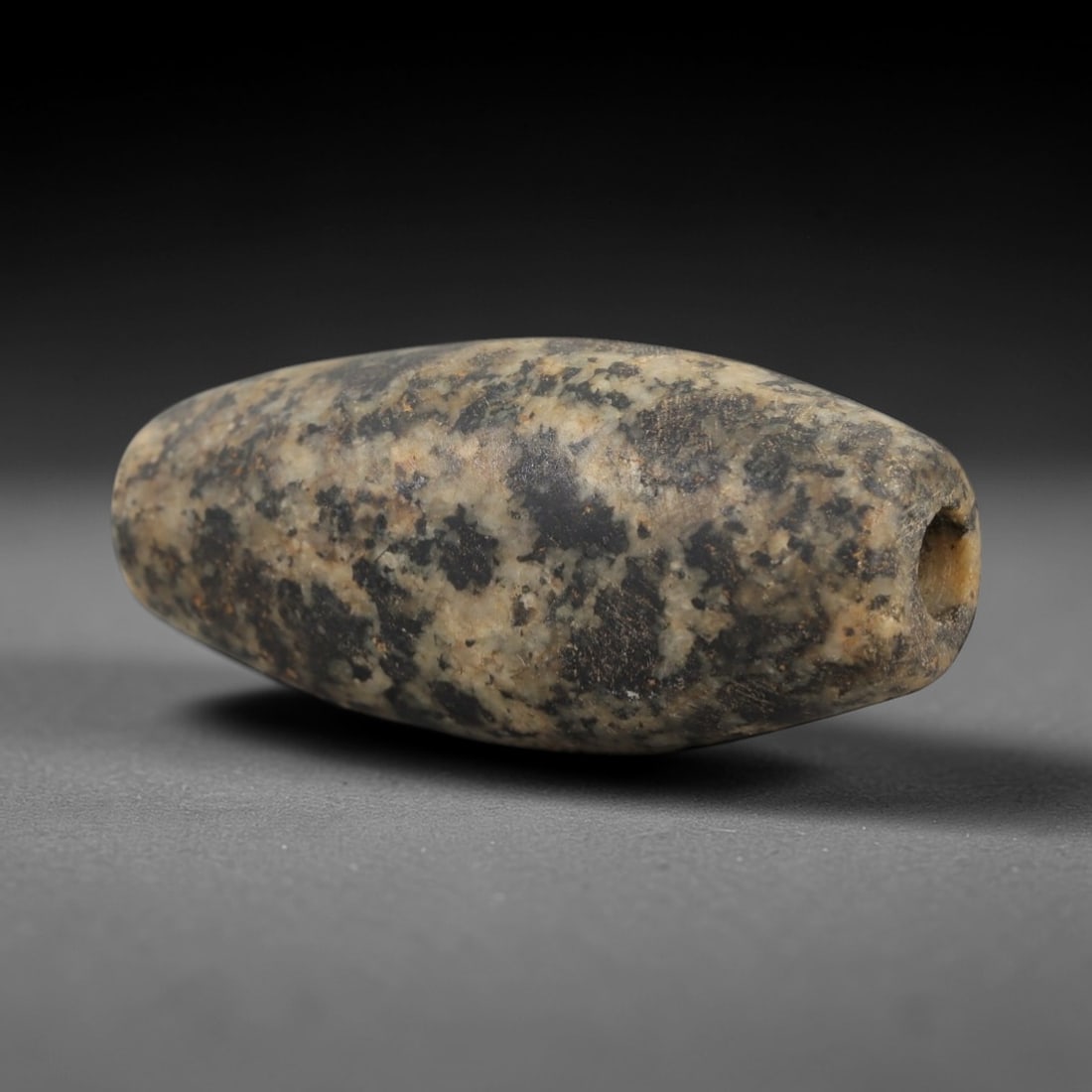 Ancient Egyptian Stone Bead - Carved Ritual Amulet, 69AE: Carved from mottled grey stone, this bead features a cylindrical form with a central perforation, typical of small Egyptian ritual beads from the Late Period. The surface displays a natural, even pati