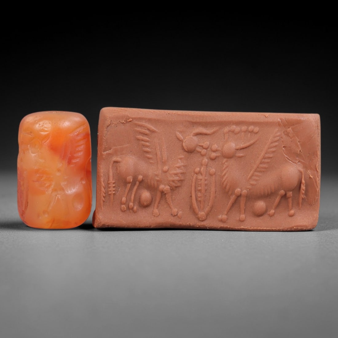 Ancient Carnelian Cylinder Seal - Near Eastern Mythical Beasts, 49AN: Carved from polished orange carnelian, this cylinder seal depicts two winged mythical beasts flanking a central sacred tree motif, with incised details defining their horns, wings, and segmented limbs
