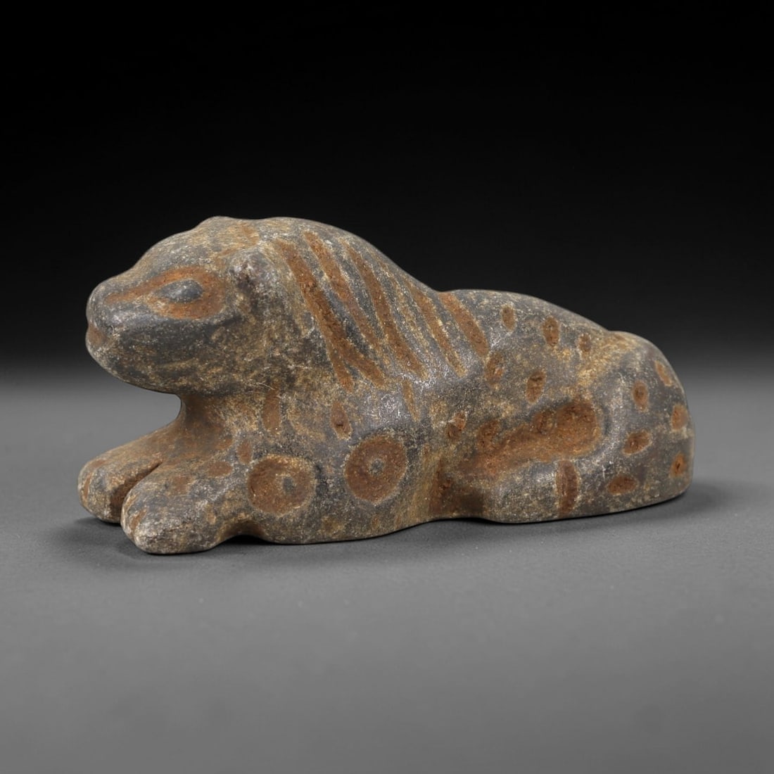 Ancient Stone Zoomorphic Amulet - Near Eastern Feline Figure, 12AM: This amulet is carved from dark stone, depicting a recumbent feline in a crouched posture, with incised linear and circular motifs defining the mane, spots, and facial features. The piece reflects the