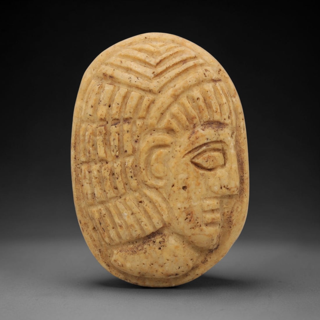 Ancient Stone Amulet - Near Eastern Bust Plaque Pendant, 26ZX: Carved from light-colored stone, the amulet is an oval plaque crafted in the ancient Near Eastern style, featuring a raised relief portrait of a human bust in profile. The surface bears incised detail