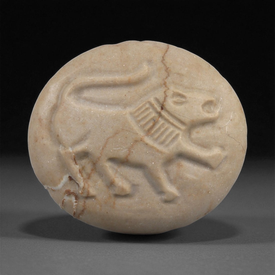 A Near Eastern Revivalist Marble Plaque with Lion, 20th Century, 35LF: Carved from light beige marble with natural veining, the plaque is shaped as an oval with a slightly convex face. The surface bears a shallow relief of a striding lion, its body outlined by broad, smo