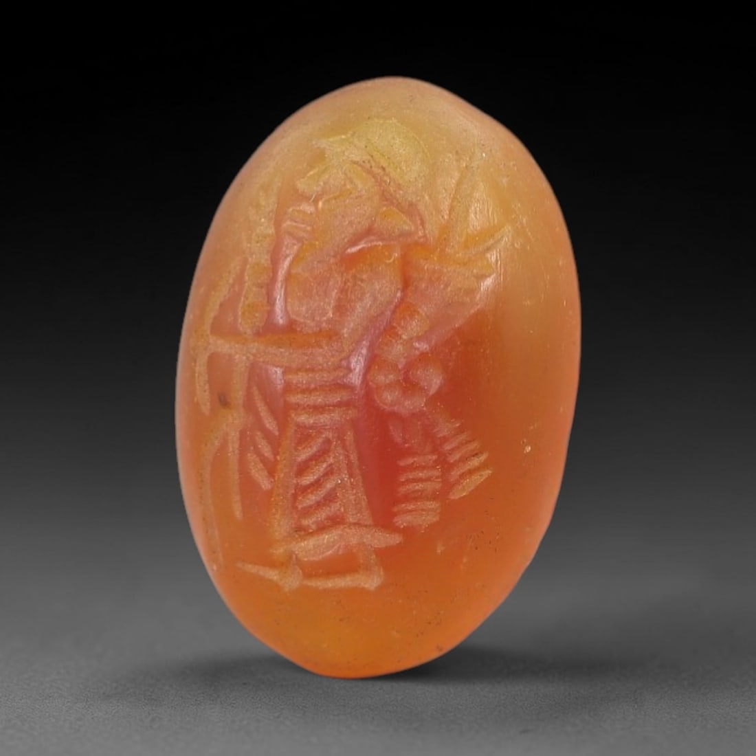 Ancient Carnelian Anthropomorphic Intaglio - Near Eastern Seal Amulet, 83AM: Carved from warm orange carnelian, this 2x1.3cm oval intaglio features a finely incised anthropomorphic figure in profile, adorned with a headdress, pleated skirt, and holding symbolic elements (likel