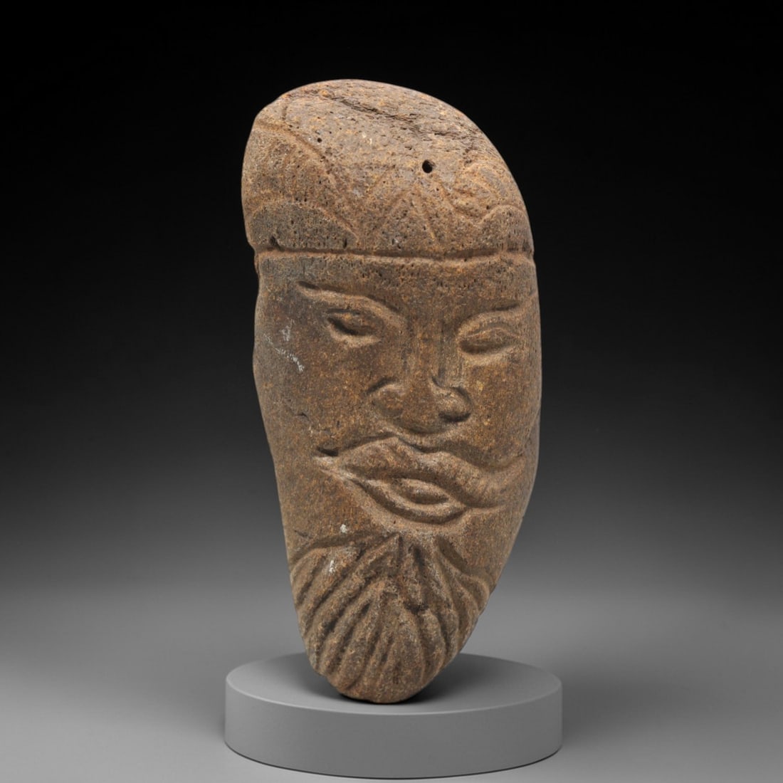 Ancient Stone Bearded Head - Near Eastern Idol, 80ZV: This artifact is a carved stone head of anthropomorphic form, executed in a granular, earth-toned stone consistent with ancient Near Eastern lithic traditions. The relief carving depicts a mature male