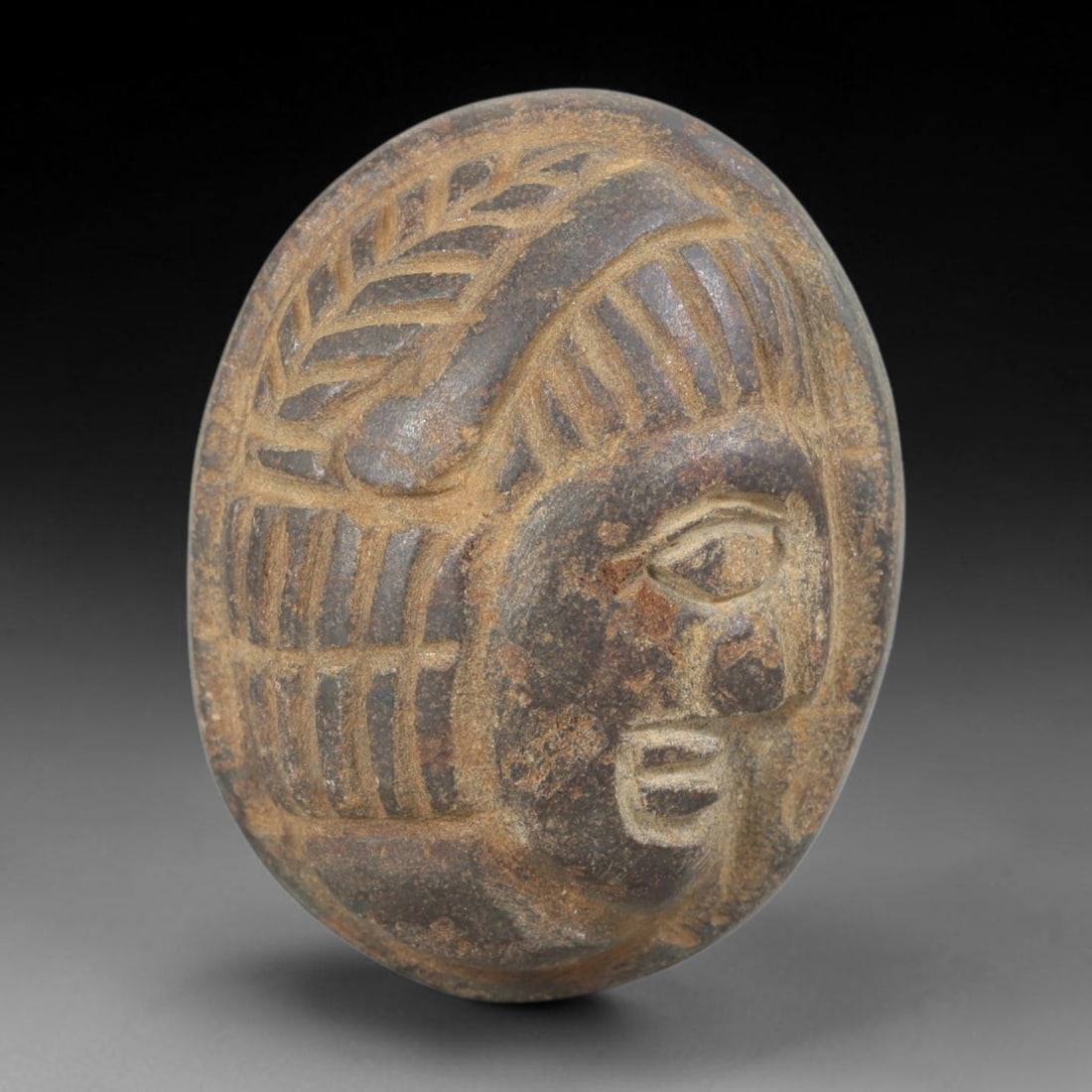 Ancient Stone Head Plaque - Near Eastern Amulet, 45FY: Carved from a dark grey stone with warm brown mineral accretions, this amulet takes the form of a stylized human head in high relief, set within an oval plaque, reflecting the iconographic traditions