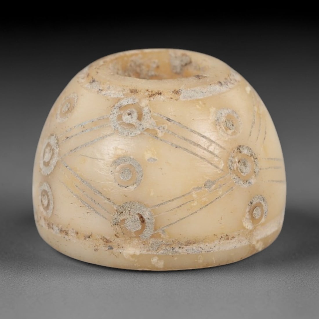 Ancient Bone Spindle Whorl - Near Eastern Radiating Lines, 62XR: This ancient Near Eastern spindle whorl is carved from bone, featuring a biconvex form with a central perforation. The surface is incised with radiating linear and circular motifs, a common decorative