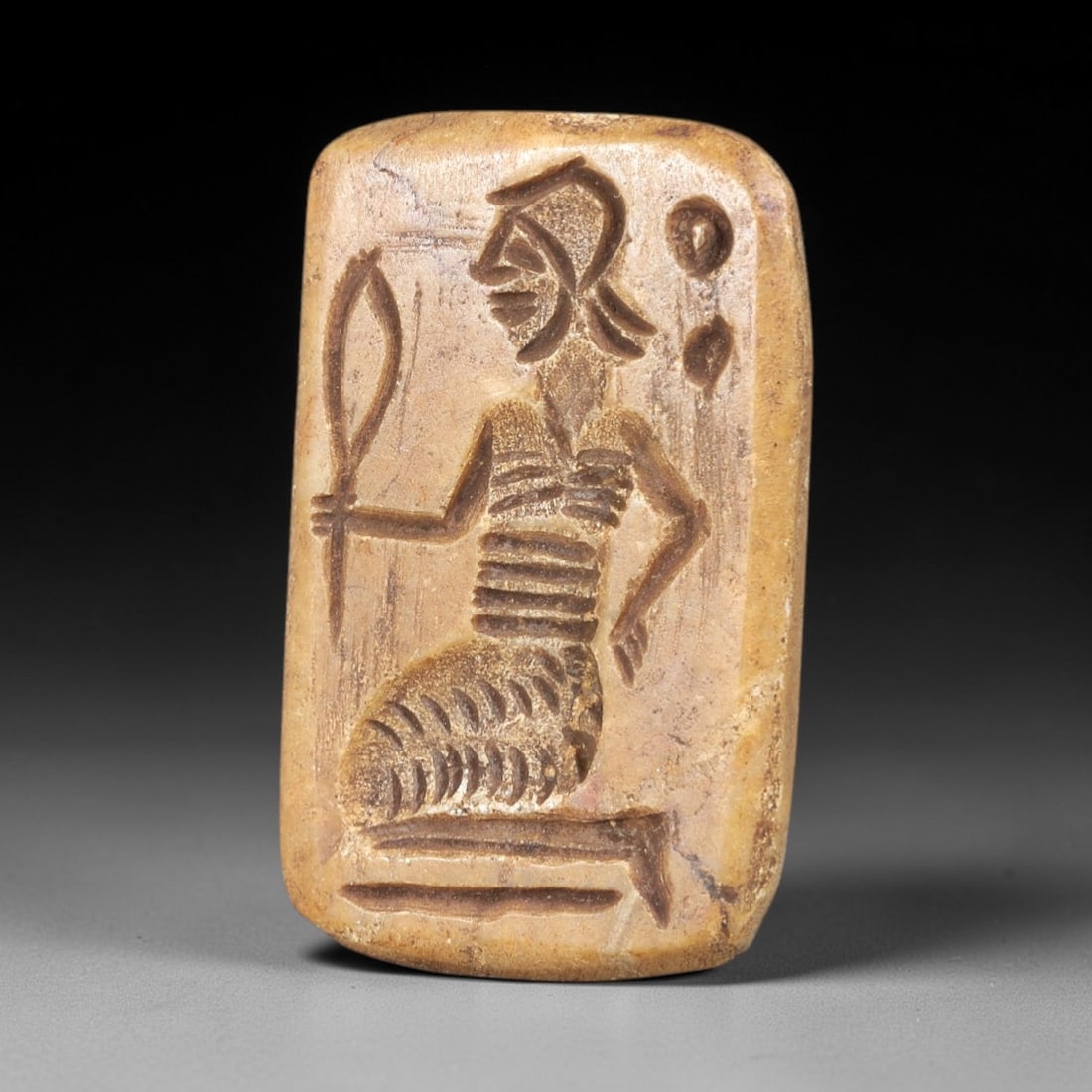 Ancient Stone Stamp Seal - Near Eastern Anthropomorphic Figurine, 52AS: This ancient Near Eastern stamp seal is carved from stone, formed as a rectangular plaque with incised decoration depicting a seated anthropomorphic figure in profile, holding a leaf-like object in on