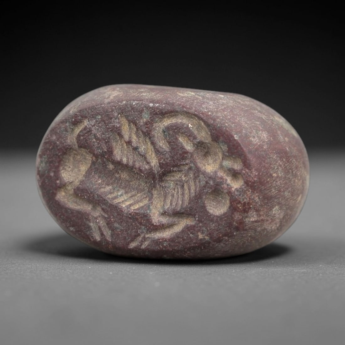 Ancient Stone Hemispherical Stamp Seal - Near Eastern Winged Creature Figure, 22AN: Carved from a dense, dark reddish-brown stone, this hemispherical stamp seal features a flat, oval face incised with a stylized winged creature in profile. The creature is depicted with a horned head,