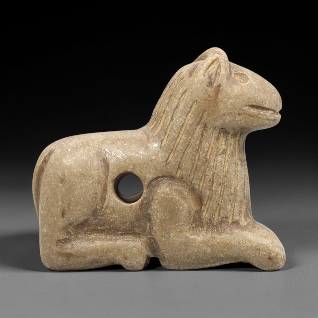 Ancient Stone Canid Amulet - Near Eastern Recumbent, 59ZR: Carved from warm beige-hued stone with a subtle matte patina and fine mineral flecks, executed in a stylized recumbent form characteristic of Near Eastern zoomorphic amulet craftsmanship. The figure d