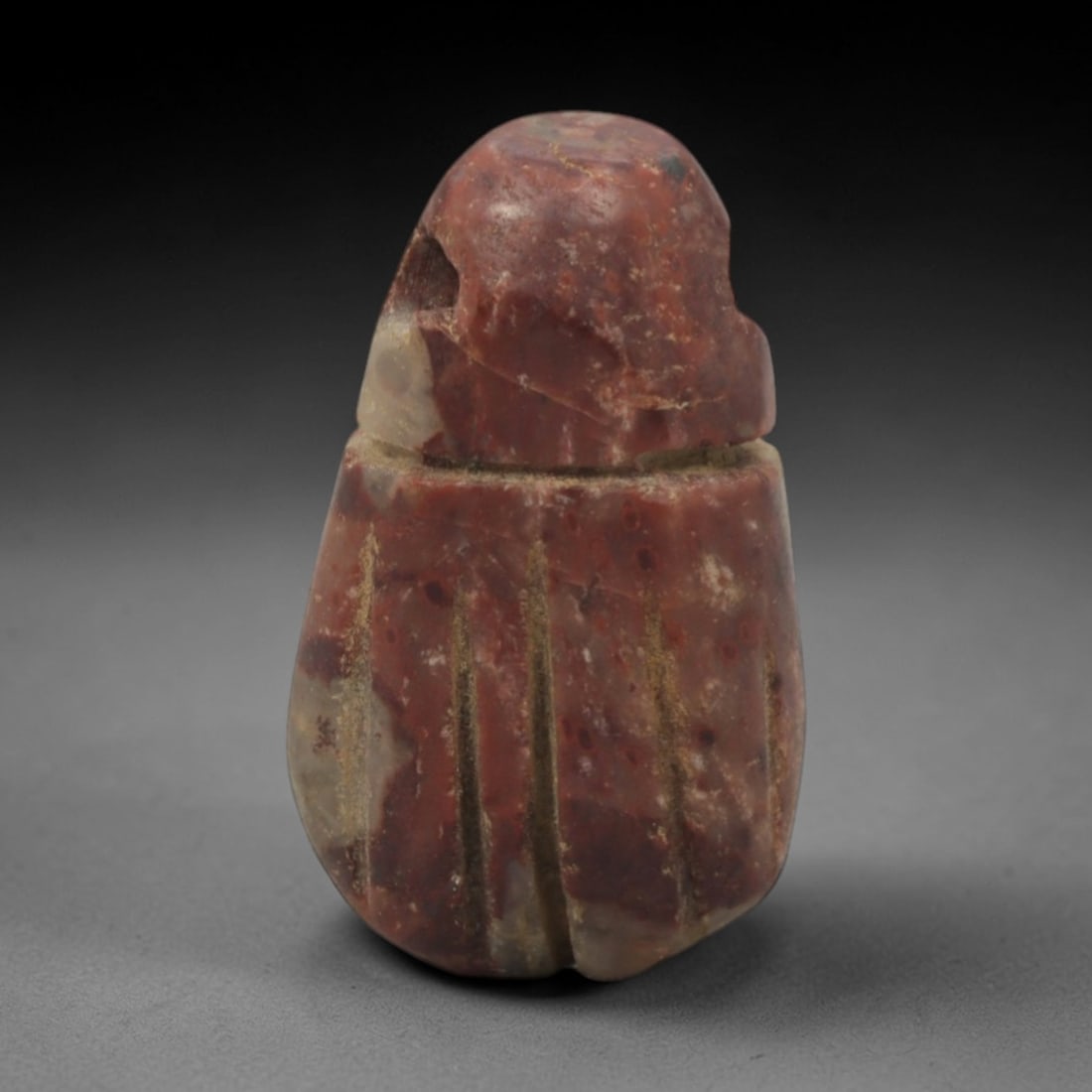 Ancient Stone Scarab - Egyptian Amulet Figurine, 3XR: Carved from rich red stone with natural veining, this amulet depicts a stylized scarab beetle with a segmented body and vertical incised lines representing the elytra. The form and iconography are cha