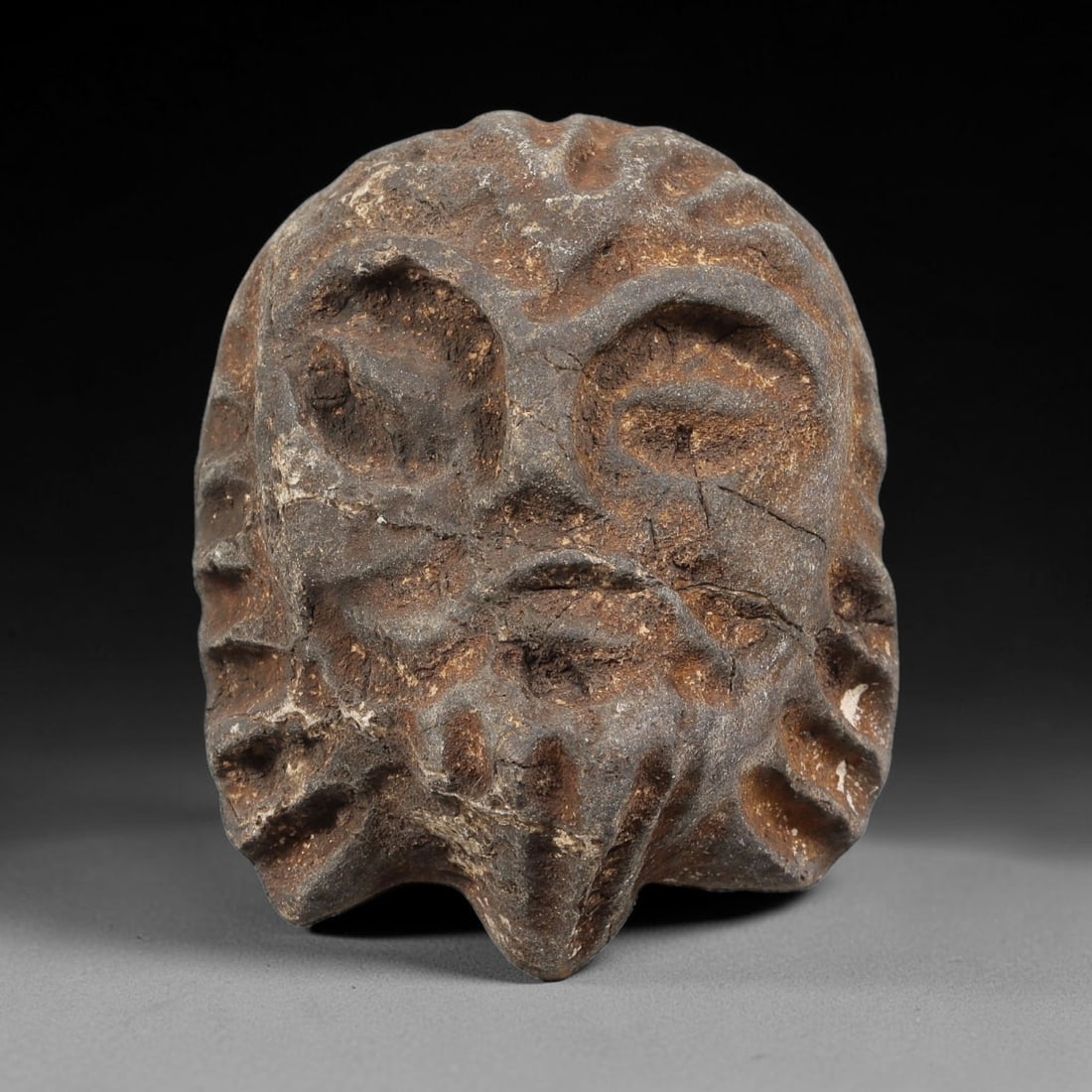 Ancient Stone Maskette - Near Eastern Anthropomorphic Head, 46WJ: Carved from a coarse, porous stone, this artifact takes the form of a stylized anthropomorphic maskette, rendered with bold, deeply incised features. The eyes are carved as large, recessed almond shap