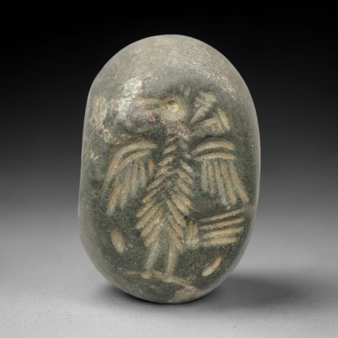 Ancient Stone Hemispherical Stamp Seal - Near Eastern Bird Figure, 89ZS: Crafted from dense, greenish stone, this hemispherical stamp seal bears incised carving of a stylized bird figure, likely an eagle or raven, with outstretched wings, segmented body detailing, and line