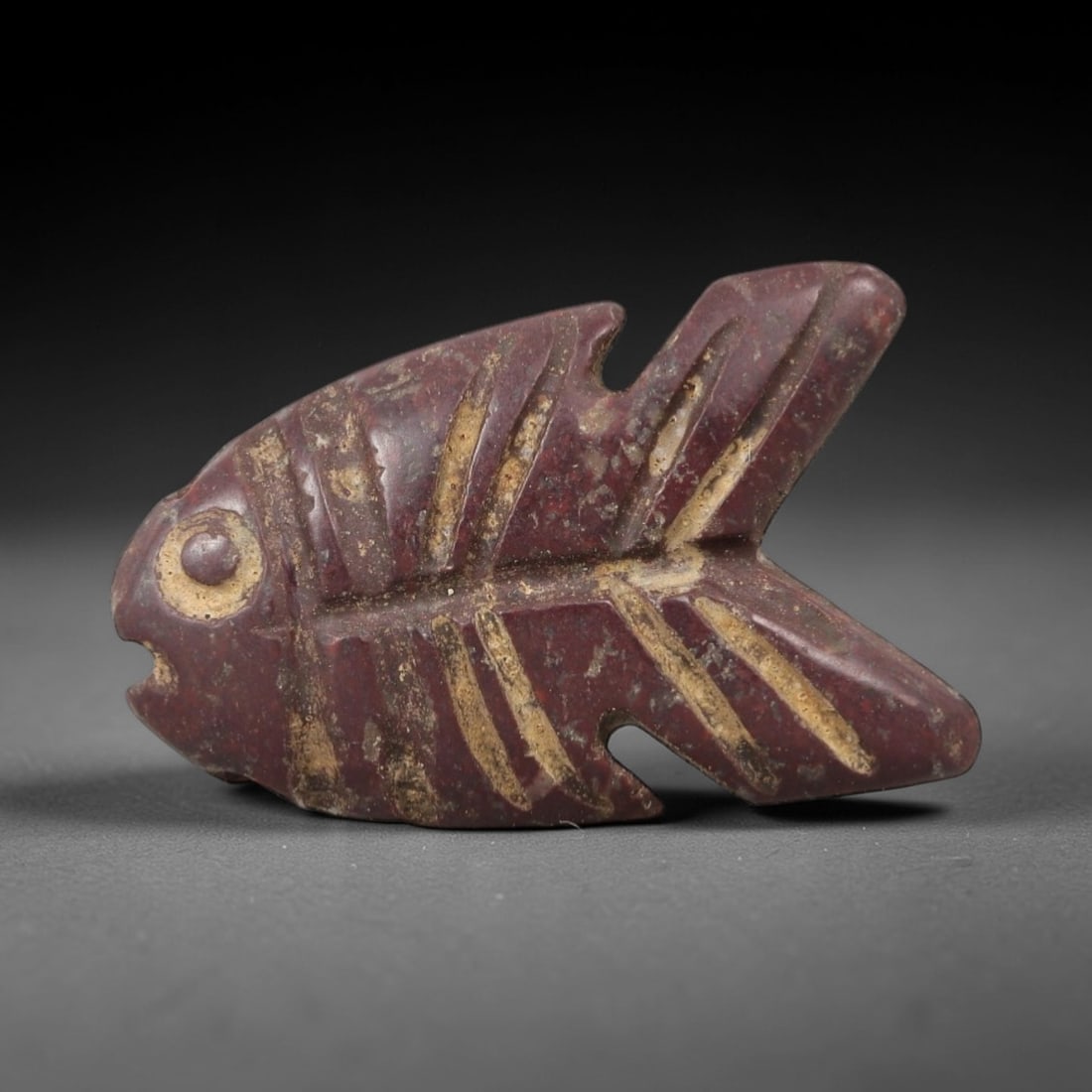 Ancient Stone Fish Amulet - Egyptian Ritual Figurine, 30AG: This amulet is carved from a single piece of deep reddish-purple stone, exhibiting a granular texture with pale ochre incrustations filling the carved lines across its surface. The form is stylized as