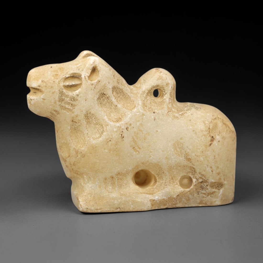 Ancient Marble Feline Amulet - Near Eastern Figurine, 59XV: Crafted from pale marble, this small amulet depicts a stylized recumbent feline in a compact form. The robust body rests on a solid base, with incised details defining the head, ears, and facial featu