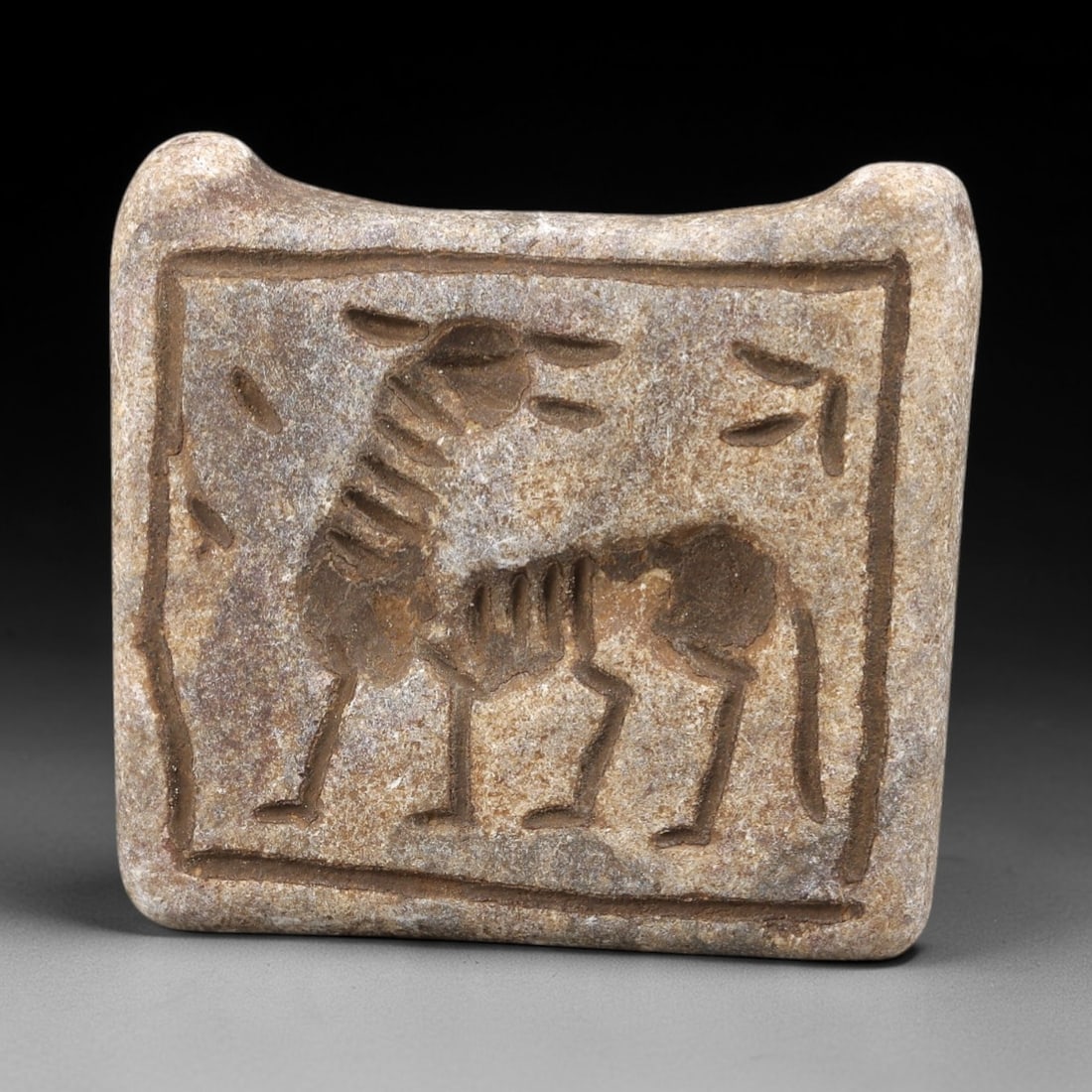 Ancient Stone Double Convex Amulet - Near Eastern Bovid, 92FX: Carved from a double-convex stone plaque, this amulet features a relief depiction of a stylized bovid, likely a bull or ram, with incised lines defining its segmented neck, ribcage, and four-legged st
