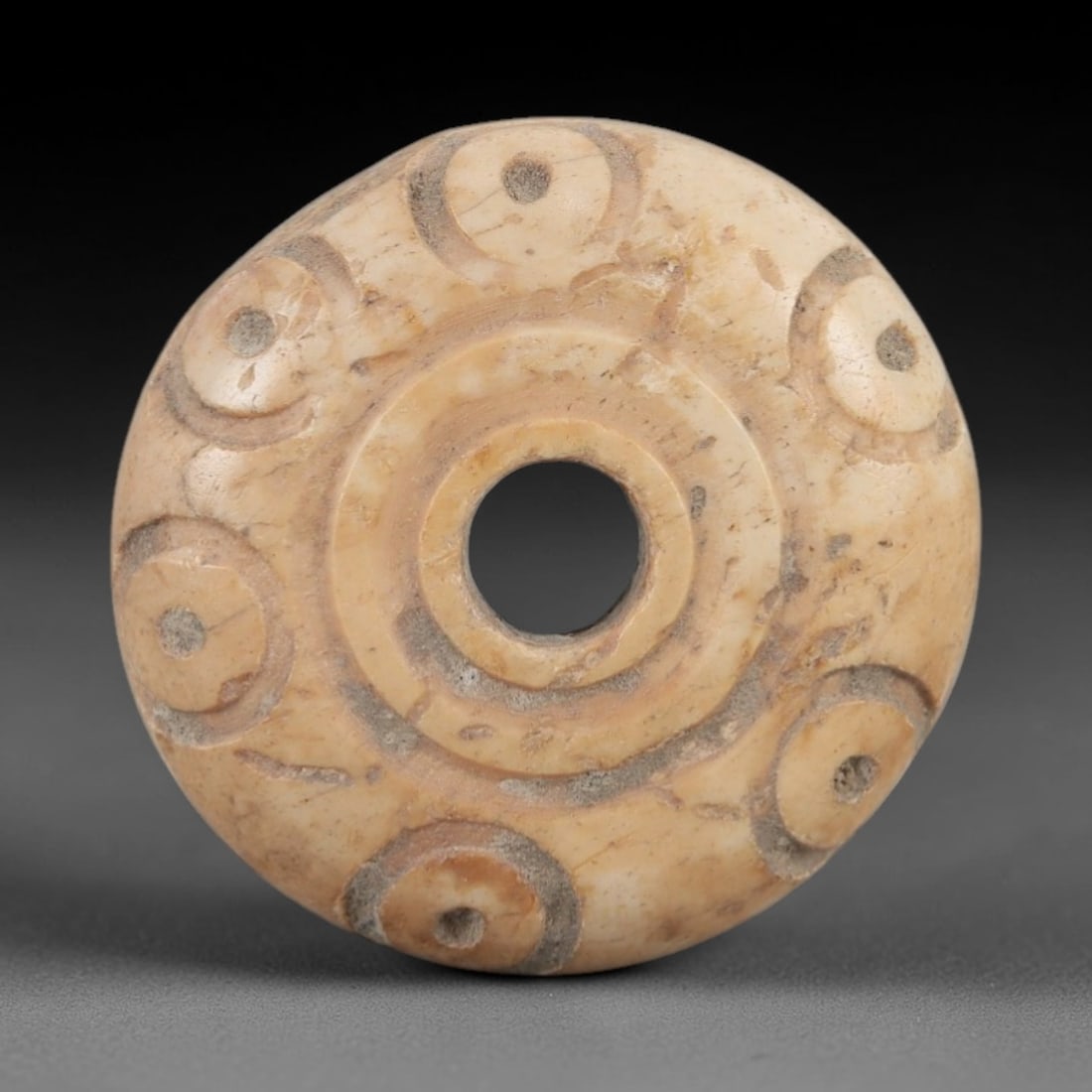 Ancient Bone Spindle Whorl - Near Eastern Dot-in-Circle Motif, 69XR: This ancient Near Eastern spindle whorl is crafted from bone, featuring incised dot-in-circle motifs encircling the central perforation. The compact, ring-like form is a traditional textile tool used