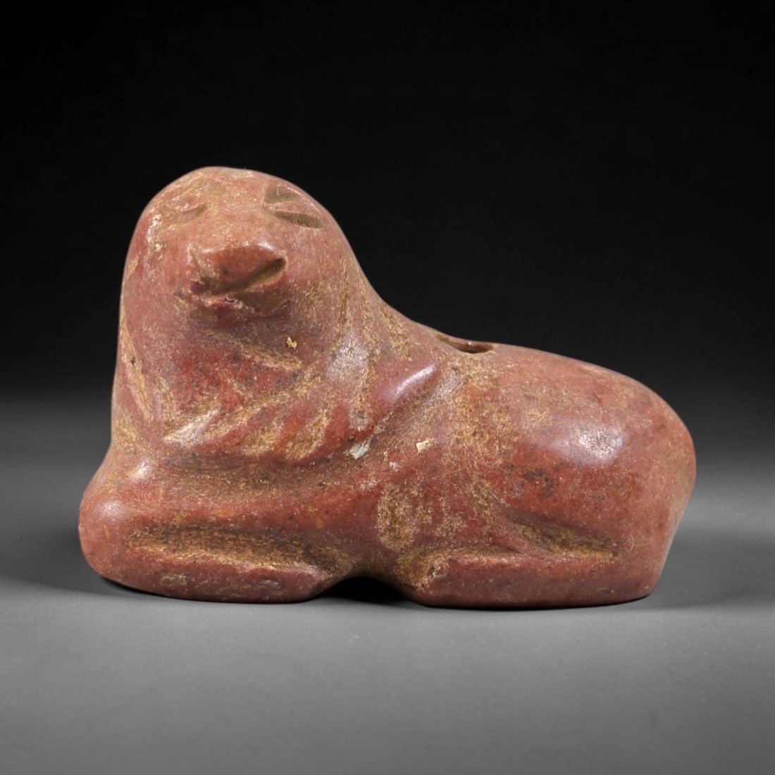Ancient Red Stone Lion - Near Eastern Amulet Figurine, 38AS: This amulet is carved from compact reddish stone, displaying a warm, earthy patina consistent with ancient Near Eastern burial contexts. The form depicts a recumbent lion, rendered in a stylized, geom