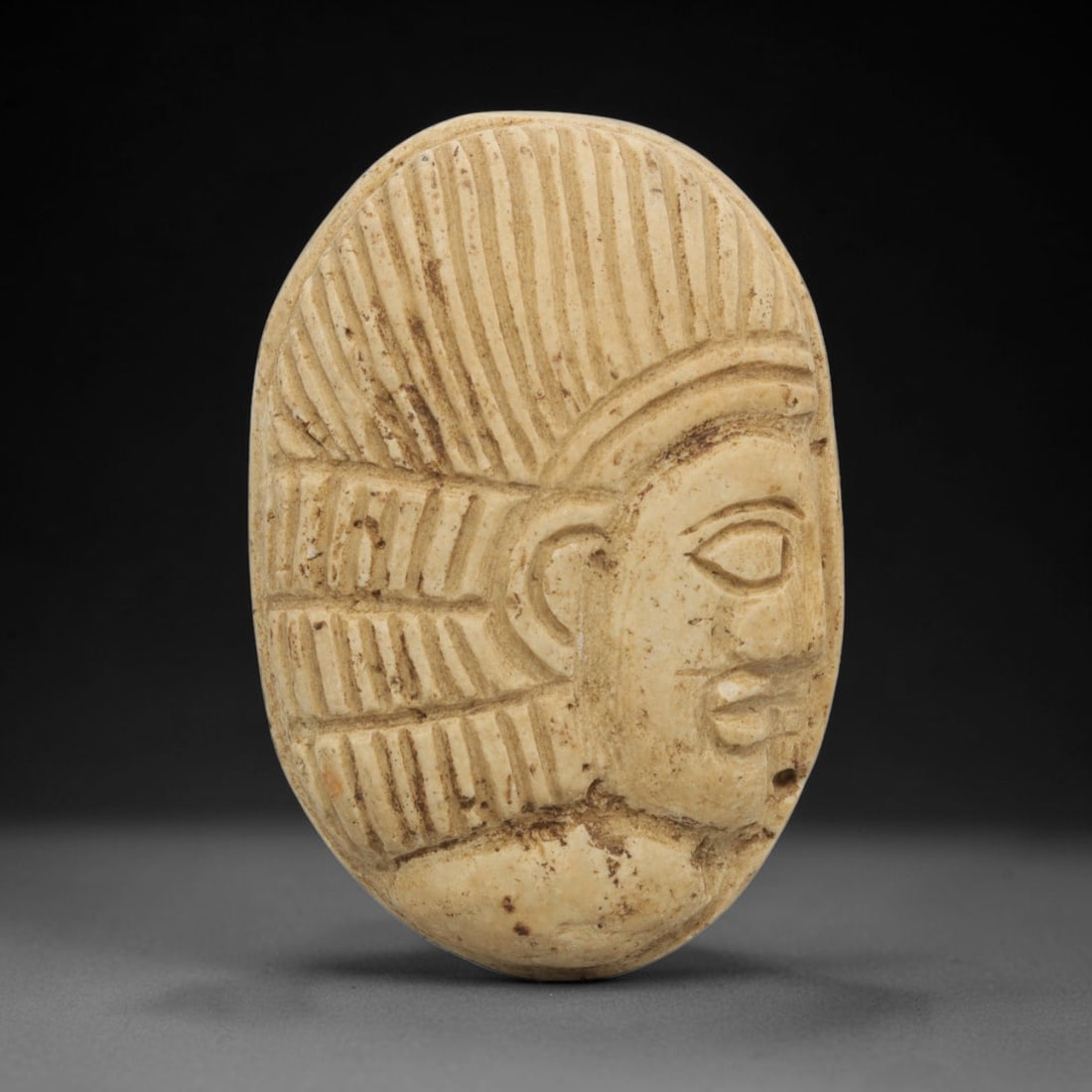 Ancient Stone Bust Amulet - Near Eastern Figurine, 92ZT: Carved from a fine-grained, creamy stone with a warm, aged patina, this oval amulet features a deeply incised relief depiction of a Near Eastern human bust in profile, with distinct stylized facial fe