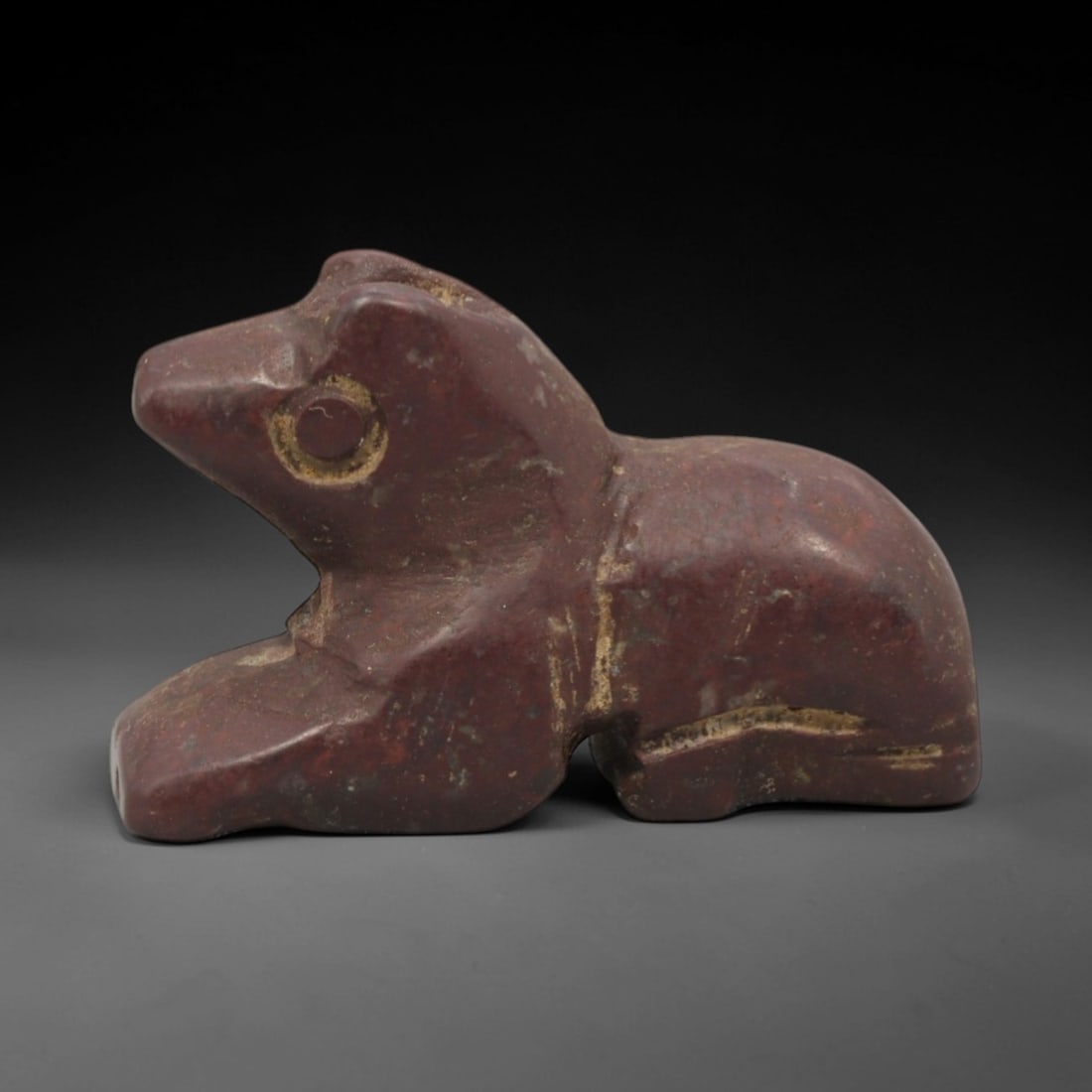 Ancient Stone Recumbent Animal - Egyptian Amulet Figurine, 9XR: Carved from a dense reddish-brown stone, this amulet depicts a recumbent zoomorphic creature, likely a feline or jackal, with a distinct circular incised eye and simplified anatomical forms typical of