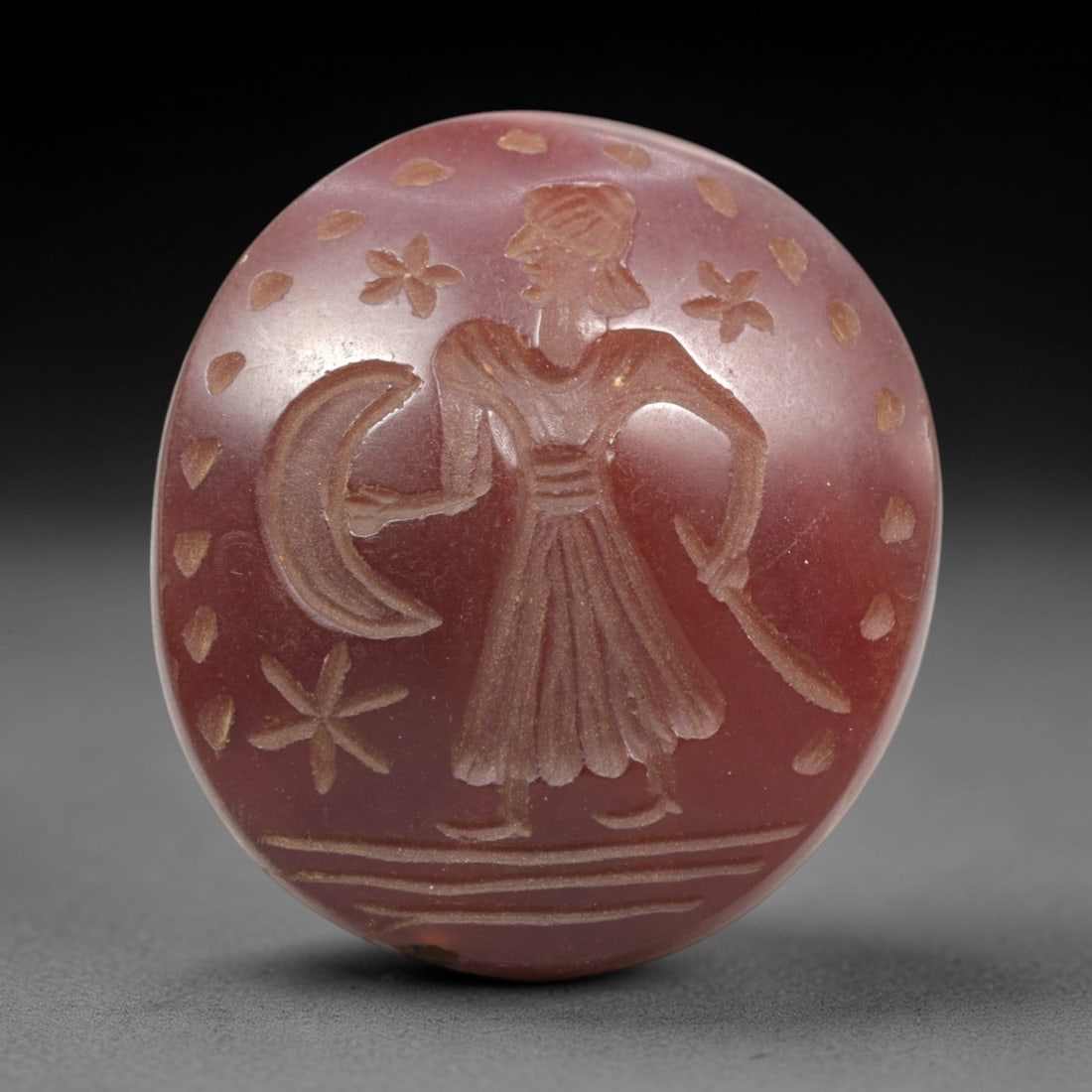 Ancient Agate Intaglio Bead - Near Eastern Celestial Figure, 75ZR: Carved from rich reddish-brown agate, this oval intaglio bead depicts a standing anthropomorphic figure in raised relief, holding a crescent moon in one hand and a blade-like object in the other, surr