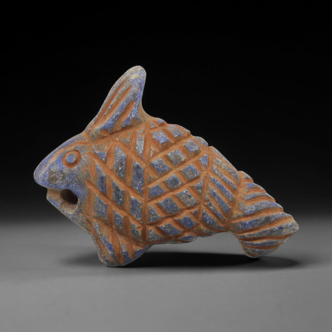 Ancient Lapis Lazuli Fish Amulet - Near Eastern Carved Figurine, 24WJ: Carved from lapis lazuli, the amulet takes the form of a stylized fish, rendered with a robust, compact silhouette. The head features a circular incised eye and an open mouth, with a suspension hole d