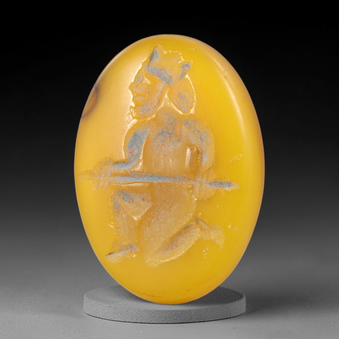 Ancient Yellow Stone Warrior Intaglio - Near Eastern Amulet, 22AR: This oval intaglio is carved from a smooth, golden-yellow stone, featuring a high-relief depiction of a kneeling warrior figure in profile, wielding a spear. The figure is adorned with a stylized head