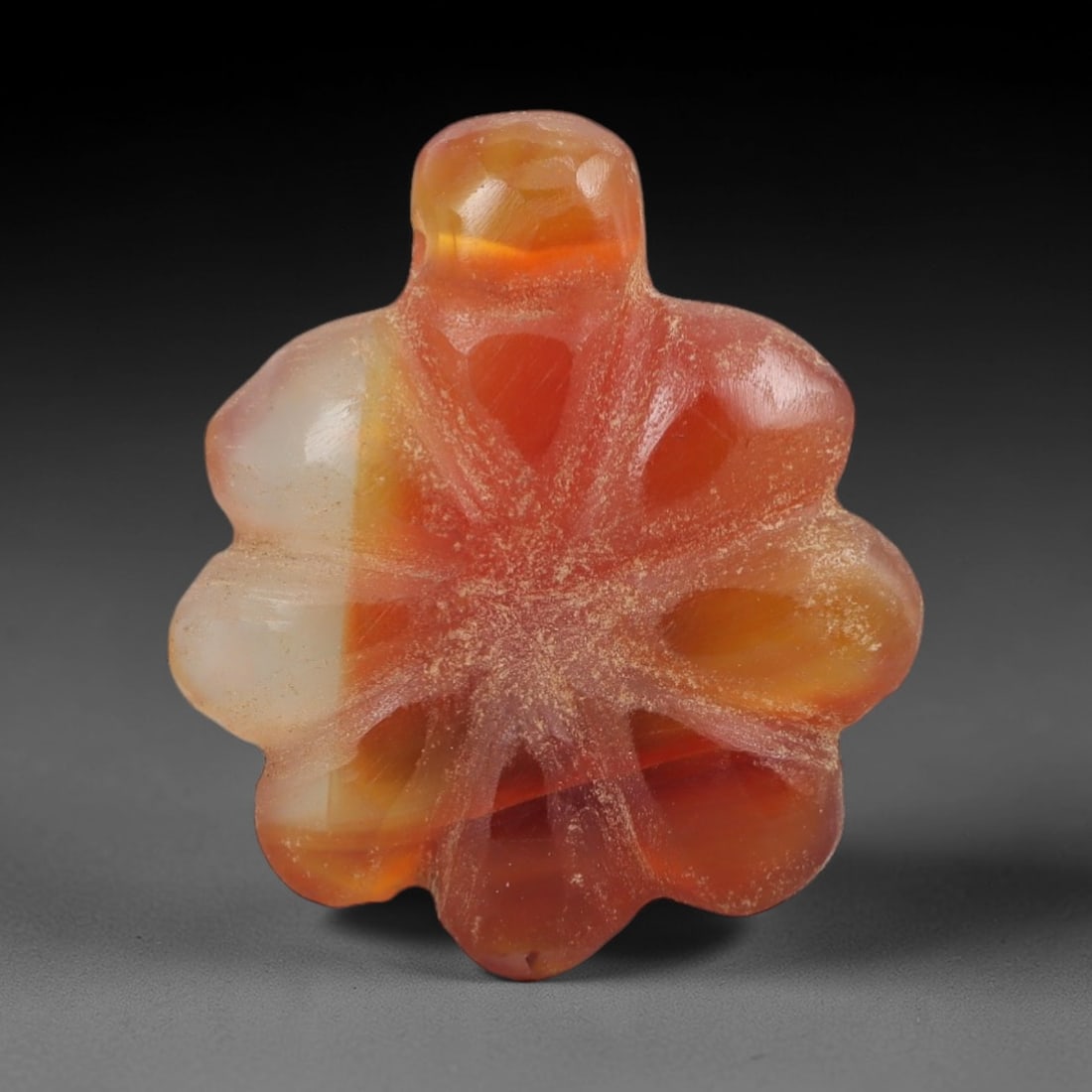 Ancient Agate Flower Amulet - Egyptian Figurine, 58AJ: Carved from banded orange-and-white agate, this amulet is formed as a stylized six-lobed flower with incised radiating linear detailing across the surface to denote petal veins, and a raised suspensio