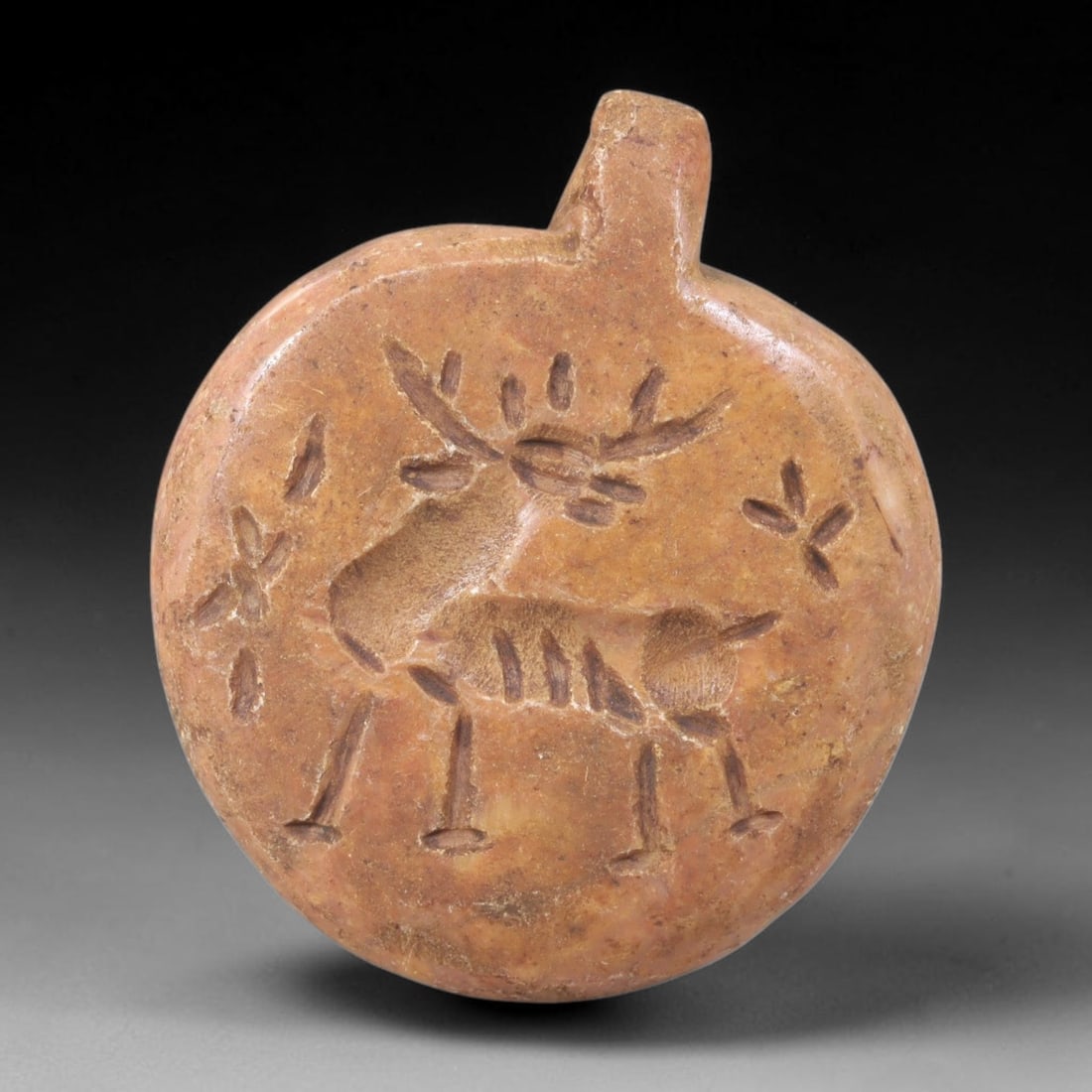 Ancient Stone Stag Amulet - Near Eastern Plaque, 31FY: Carved from a warm tan limestone, this amulet takes the form of a circular plaque with a small suspension lug at the top, characteristic of ancient Near Eastern amuletic and glyptic traditions. The pl