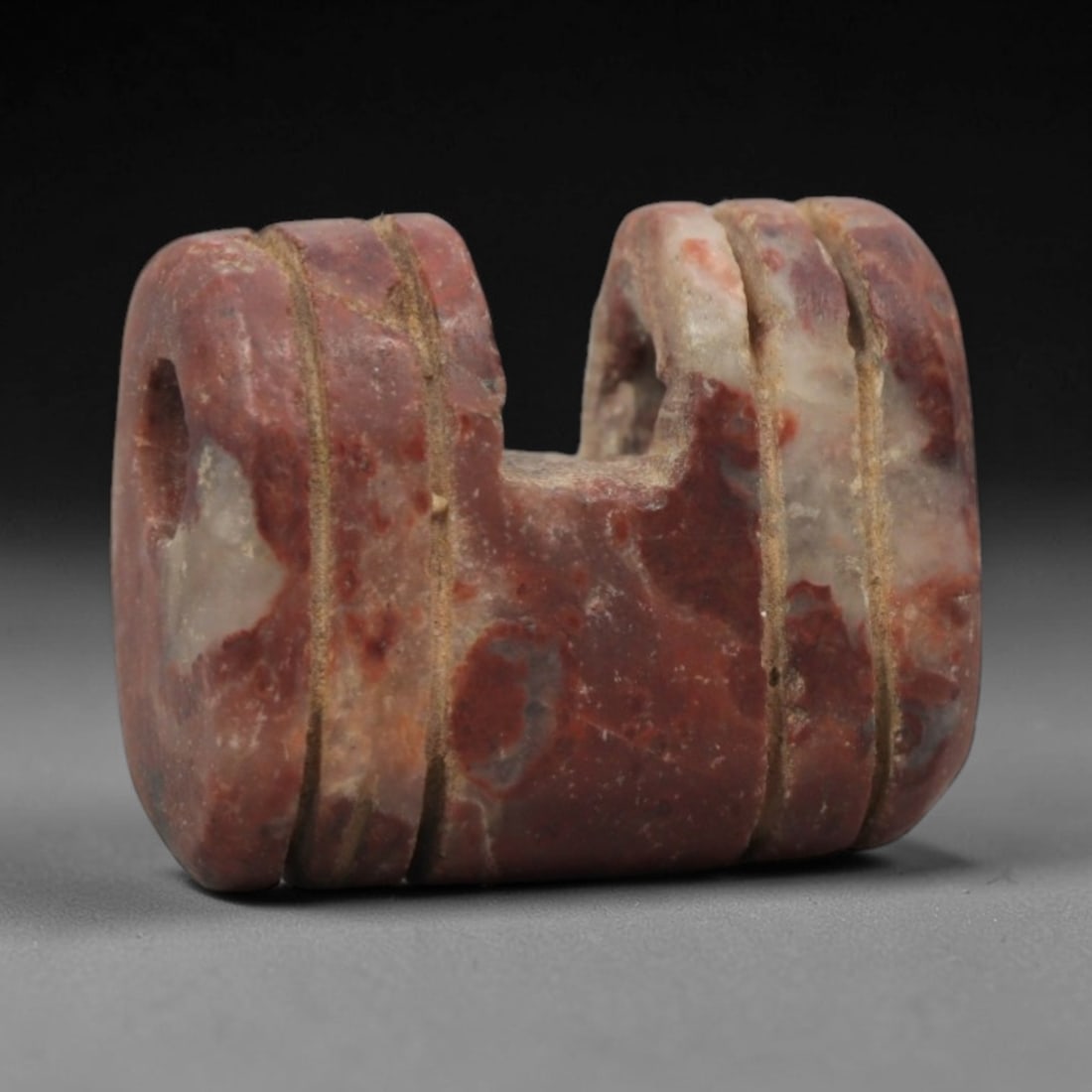 Ancient Near Eastern Red Stone Double Amulet - Carved Bead, 52ZV: This amulet is carved from a rich, reddish-brown stone with natural lighter veining, likely brecciated jasper or red steatite, featuring a symmetrical double-form design. The piece is composed of two