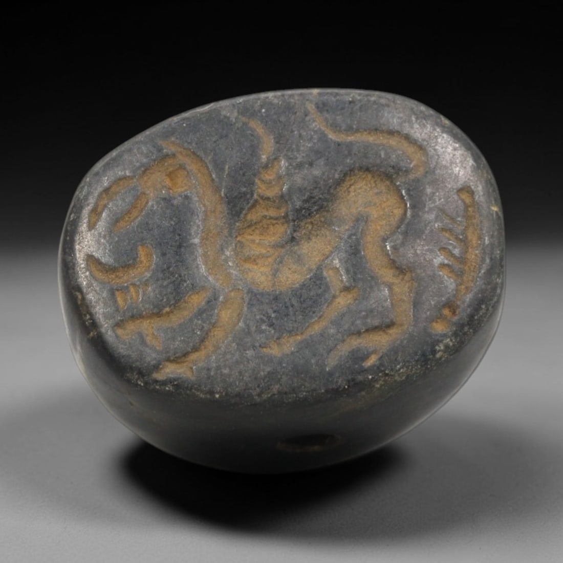 Ancient Stone Stamp Seal - Near Eastern Caprine Figure, 66ZR: Carved from a dense, dark stone, this hemispherical stamp seal features a raised relief depiction of a horned caprine (likely ibex or gazelle) in dynamic posture, alongside stylized linear motifs cons
