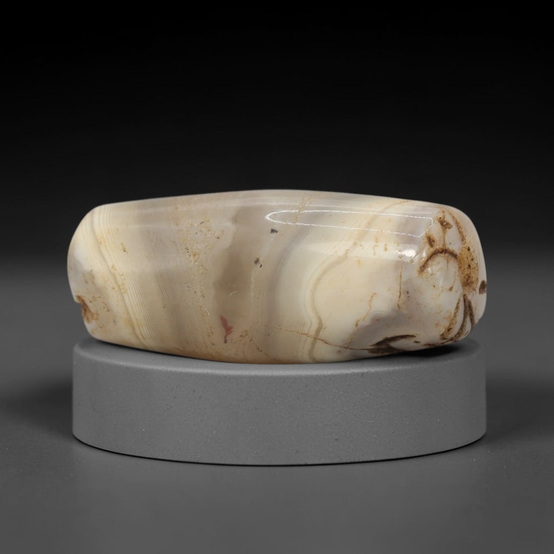 Ancient Agate Bead - Indus Valley Banded Carving: This artifact is crafted from banded agate, showcasing soft, flowing concentric banding in creamy ivory, pale grey, and warm honey tones across its polished, elongated surface. The bead is shaped in a