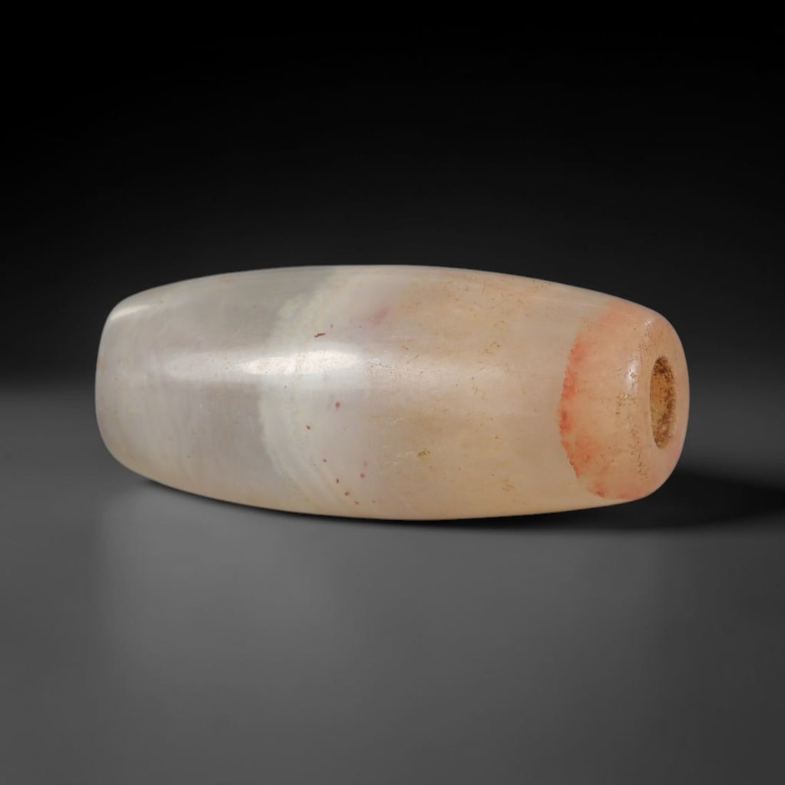 Ancient Agate Bead - Indus Valley Silk Road: Carved from banded agate, the bead is fashioned in a cylindrical barrel form with a central perforation for suspension. The stone displays natural, subtle concentric banding in creamy beige, transluce