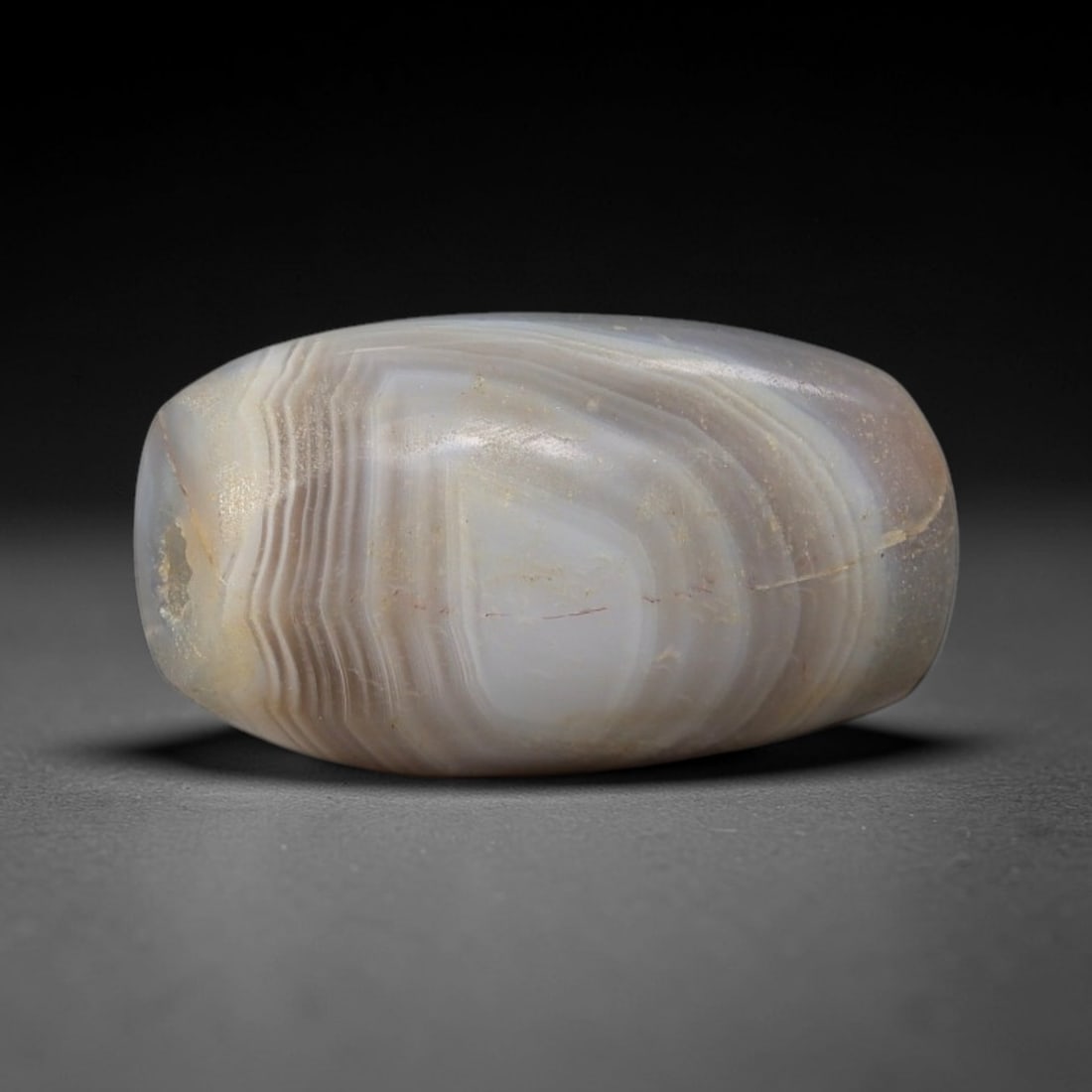 Ancient Banded Agate Bead - Indus Valley Silk Road Miniature Bead: Carved from translucent banded agate, the bead is fashioned in a compact barrel form, with a central drilled perforation for suspension. The stone exhibits fine, wavy natural banding in milky white, p