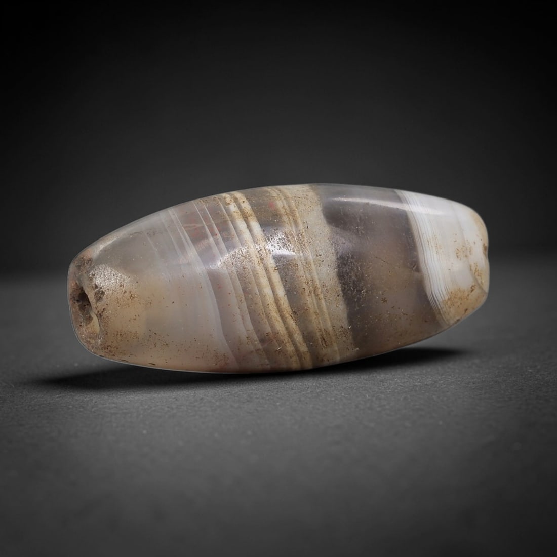 Ancient Agate Bead - Indus Valley Amulet: This artifact is crafted from banded agate, showcasing distinct concentric layers of pale grey, creamy white, and soft amber hues with subtle mineral veining, shaped into a smooth elongated barrel for